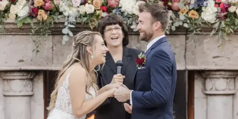 A professional wedding officiant presiding over a ceremony between a bride and groom in front of a floral arch.