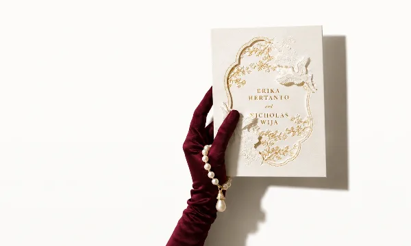 A hand wearing a burgundy velvet glove holds a cream-colored wedding invitation featuring gold foil floral patterns and the names Erika Hertanto and Nicholas Wija.