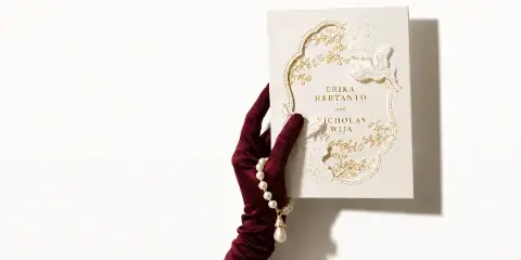 A hand wearing a burgundy velvet glove holds a cream-colored wedding invitation featuring gold foil floral patterns and the names Erika Hertanto and Nicholas Wija.