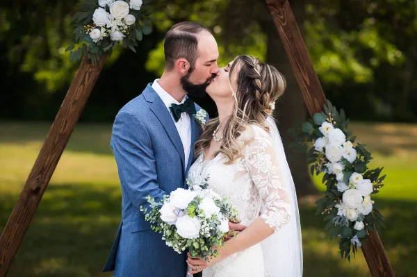 A bride and groom share a kiss under a wooden arch decorated with white flowers and greenery.