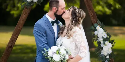 A bride and groom share a kiss under a wooden arch decorated with white flowers and greenery.