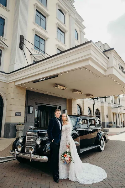 Bride and groom in formal wedding attire standing beside a vintage black car in front of a grand hotel entrance.