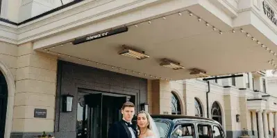 Bride and groom in formal wedding attire standing beside a vintage black car in front of a grand hotel entrance.