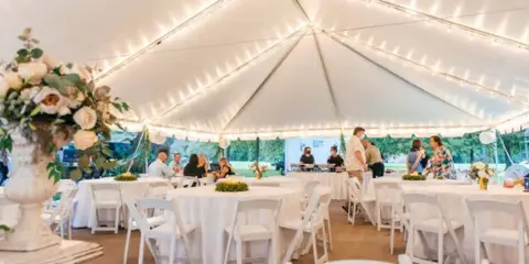Elegant wedding reception under a white tent with string lights, round tables, and white folding chairs