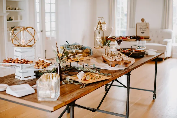 A long wooden catering table set with assorted appetizers, skewers, and decorative centerpieces in a bright indoor event space.