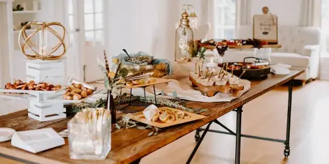 A long wooden catering table set with assorted appetizers, skewers, and decorative centerpieces in a bright indoor event space.