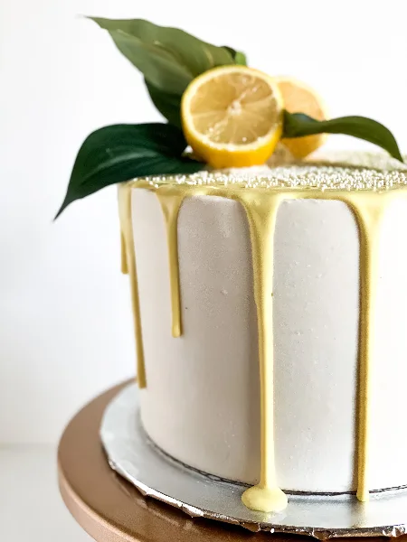 A white frosted cake with yellow drip glaze topped with lemon slices and green leaves.