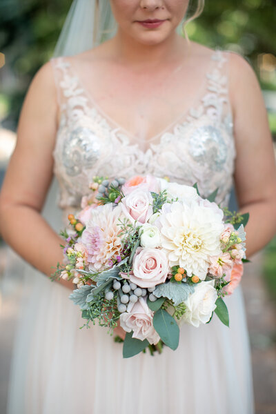 A close-up of a bride holding a custom wedding bouquet featuring pink roses, white dahlias, and greenery.