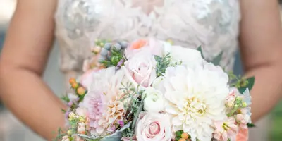 A close-up of a bride holding a custom wedding bouquet featuring pink roses, white dahlias, and greenery.
