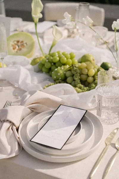 A close-up of a luxury event table setting featuring white plates, gold cutlery, a menu card, and a centerpiece of green grapes and calla lilies.