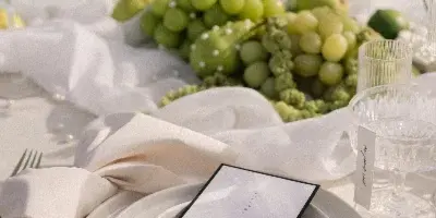 A close-up of a luxury event table setting featuring white plates, gold cutlery, a menu card, and a centerpiece of green grapes and calla lilies.