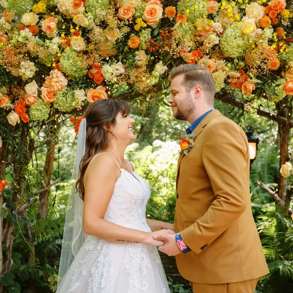 A bride and groom holding hands under a large floral archway in a garden setting.
