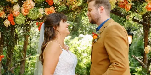 A bride and groom holding hands under a large floral archway in a garden setting.