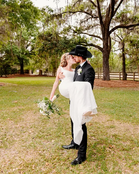 A bride in a white dress and a groom in a black suit and hat kissing while he holds her in a grassy field with trees.