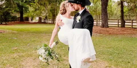 A bride in a white dress and a groom in a black suit and hat kissing while he holds her in a grassy field with trees.