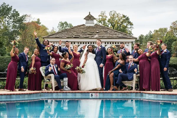A wedding party consisting of a bride, groom, and numerous bridesmaids and groomsmen posing together outdoors in front of a gazebo and pool.