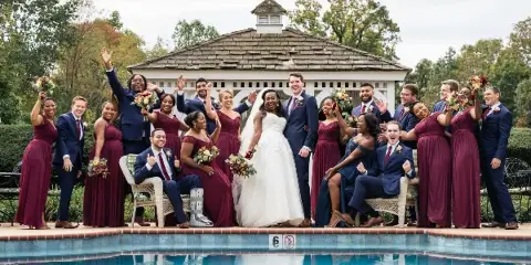 A wedding party consisting of a bride, groom, and numerous bridesmaids and groomsmen posing together outdoors in front of a gazebo and pool.
