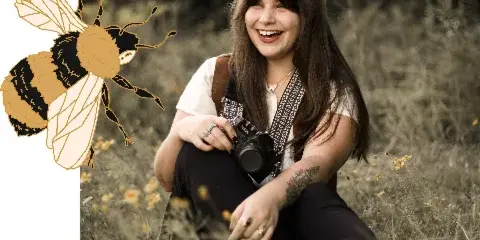 A smiling woman sitting in a field of dry grass holding a professional camera.