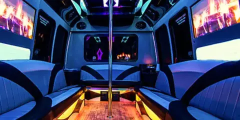Interior view of a party bus with blue and yellow LED lighting, leather bench seating, a central dance pole, and flat screen televisions.