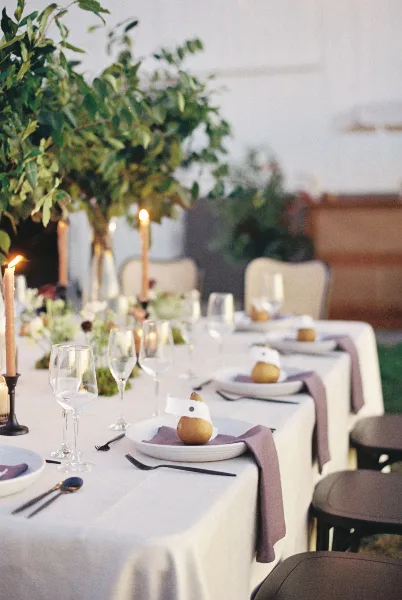 A professionally designed wedding table setting featuring a white tablecloth, tall floral arrangement, taper candles, and place settings with napkins.