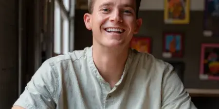 A smiling man in a light green shirt sitting at a table holding a white coffee cup.