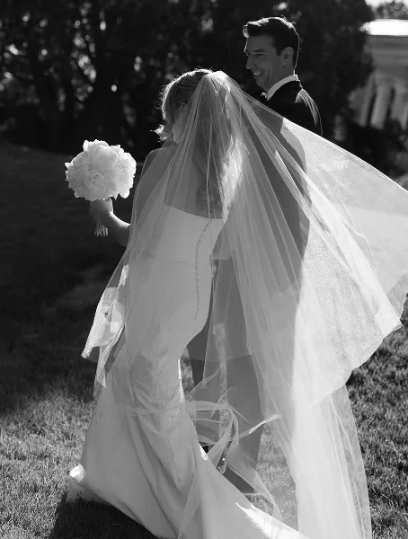 Black and white photograph of a bride and groom standing outdoors, with the bride's veil blowing in the wind.
