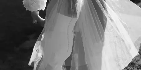 Black and white photograph of a bride and groom standing outdoors, with the bride's veil blowing in the wind.