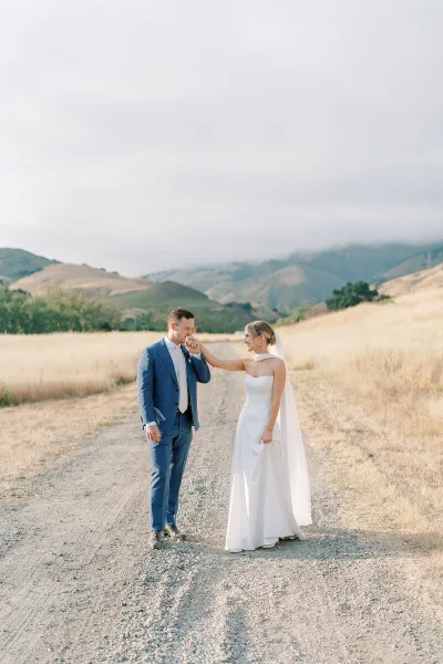 A bride and groom standing on a dirt path in a golden field with rolling hills in the background.