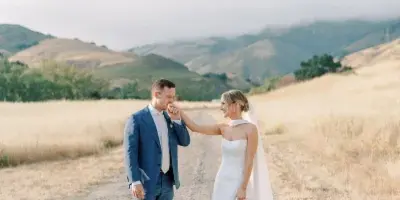 A bride and groom standing on a dirt path in a golden field with rolling hills in the background.