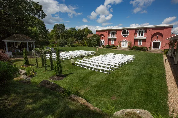 Outdoor wedding ceremony setup with white chairs on a lawn in front of a pink estate building with a gazebo and gardens.