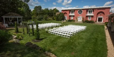 Outdoor wedding ceremony setup with white chairs on a lawn in front of a pink estate building with a gazebo and gardens.