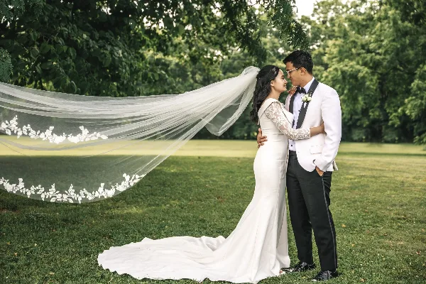 A bride and groom standing on a grassy lawn with the bride's long veil blowing in the wind.