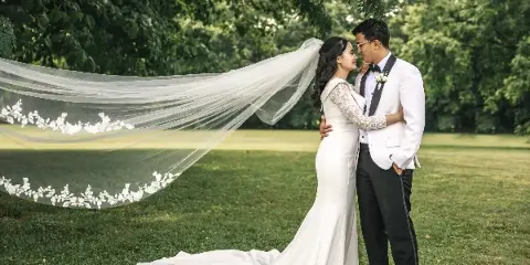 A bride and groom standing on a grassy lawn with the bride's long veil blowing in the wind.