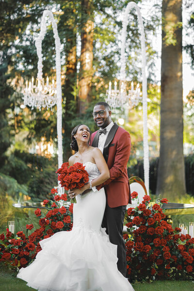 A bride in a white gown holding red roses and a groom in a maroon suit standing together outdoors near a floral arch.