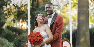 A bride in a white gown holding red roses and a groom in a maroon suit standing together outdoors near a floral arch.