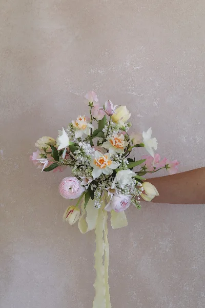 A hand holding a pastel-colored wedding bouquet with ranunculus, tulips, and white filler flowers against a textured beige wall.