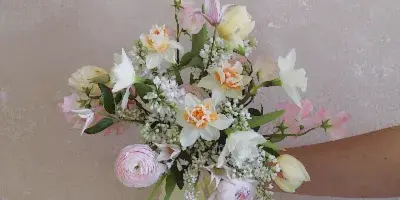 A hand holding a pastel-colored wedding bouquet with ranunculus, tulips, and white filler flowers against a textured beige wall.