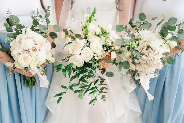 A close-up photograph of a bride and bridesmaids holding white and peach floral bouquets with greenery.