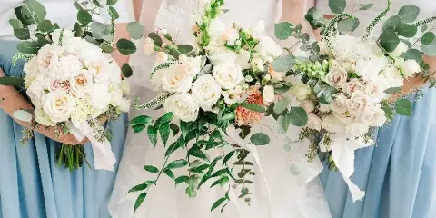 A close-up photograph of a bride and bridesmaids holding white and peach floral bouquets with greenery.