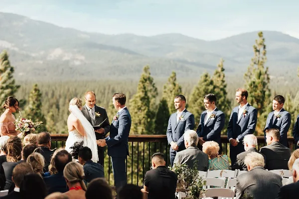 A wedding ceremony taking place on an outdoor wooden deck with a view of forested mountains in the background.