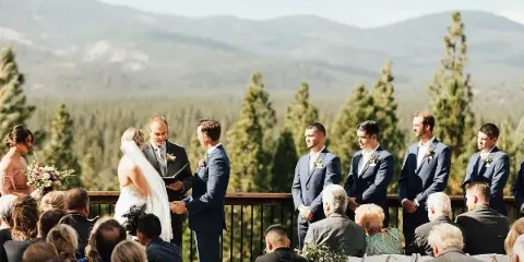 A wedding ceremony taking place on an outdoor wooden deck with a view of forested mountains in the background.