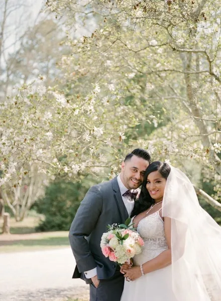 A bride and groom posing together outdoors under blooming trees, with the bride holding a floral bouquet.