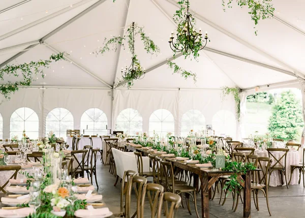 A long banquet table set with white linens, glassware, and floral centerpieces inside a white event tent.