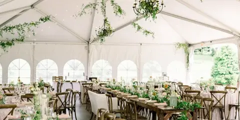 A long banquet table set with white linens, glassware, and floral centerpieces inside a white event tent.