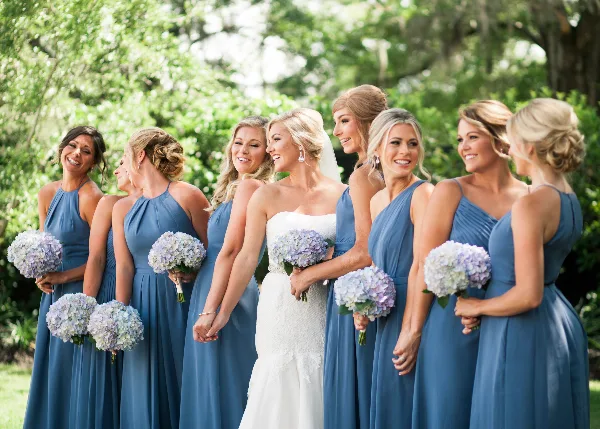 A bride in a white lace gown standing with a group of bridesmaids in blue dresses, all holding hydrangea bouquets outdoors.