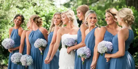 A bride in a white lace gown standing with a group of bridesmaids in blue dresses, all holding hydrangea bouquets outdoors.