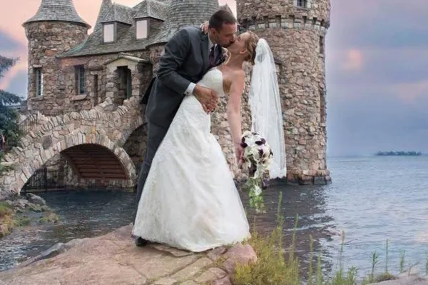 A bride and groom sharing a kiss on a rocky shore next to a stone castle-like structure.
