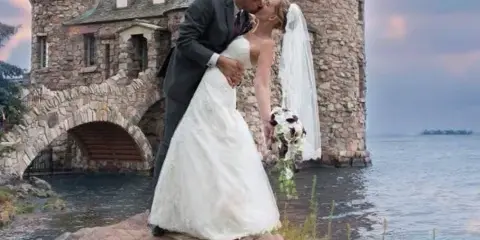 A bride and groom sharing a kiss on a rocky shore next to a stone castle-like structure.
