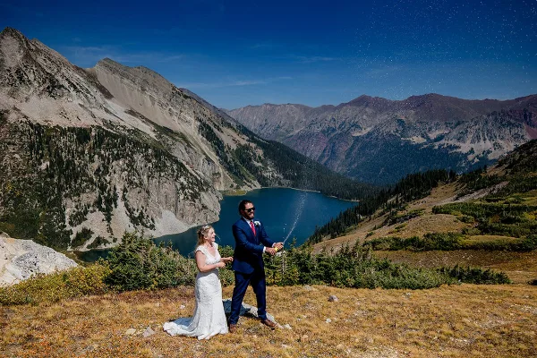 A bride and groom spraying champagne on a mountain overlook with a blue alpine lake in the background.