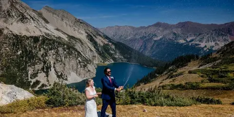 A bride and groom spraying champagne on a mountain overlook with a blue alpine lake in the background.